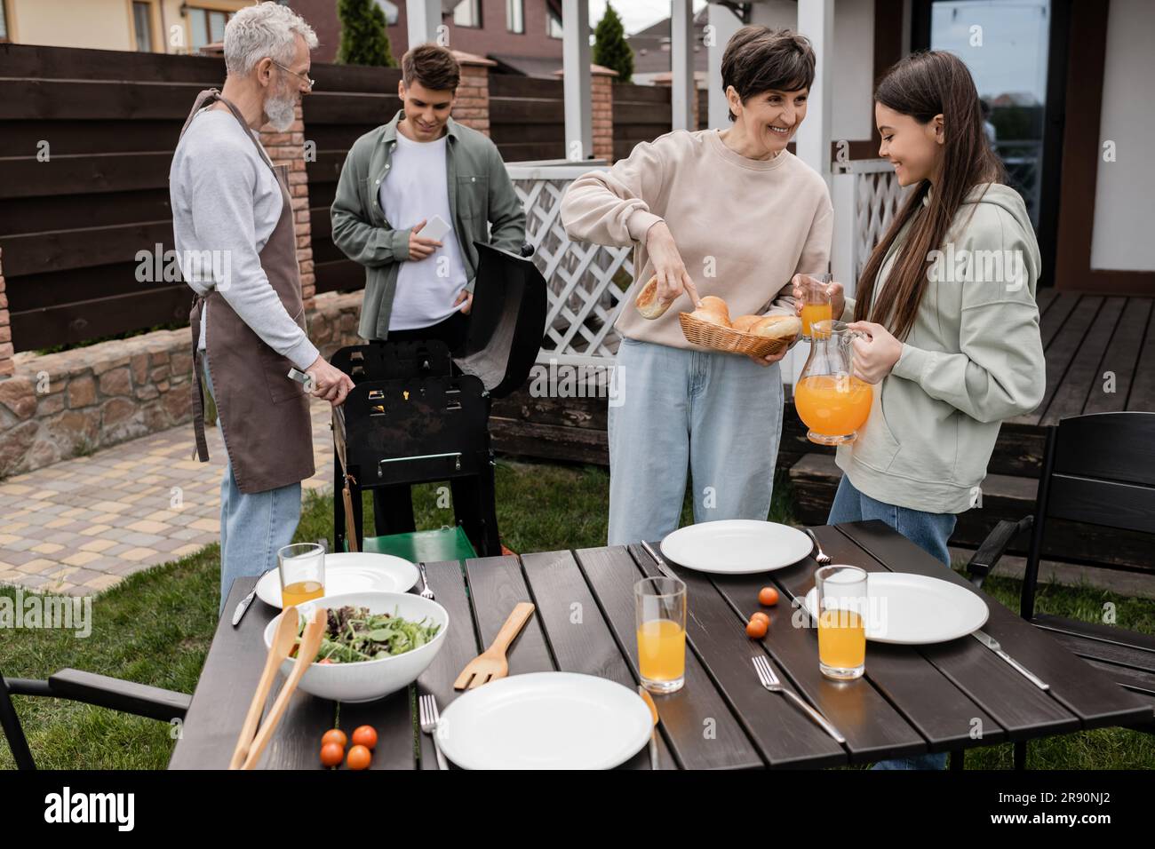 celebration of parents` day, middle aged mother showing buns to teenage daughter with jug of orange juice, father and son preparing food on bbq grill, Stock Photo